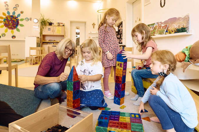 Im Kindergarten sitzt eine Frau mit mehreren Kindern auf dem Boden, die gemeinsam bunte Bauklötze stapeln.