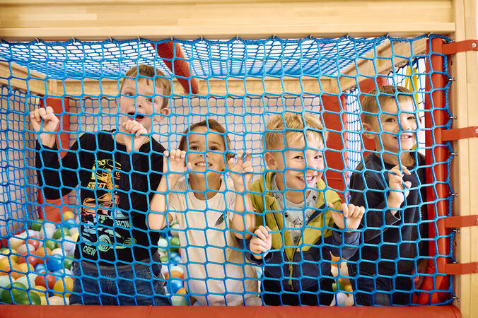 Vier Kinder schauen lächelnd durch ein blaues Netz von einem Spielbereich im Kindergarten mit bunten Bällen.