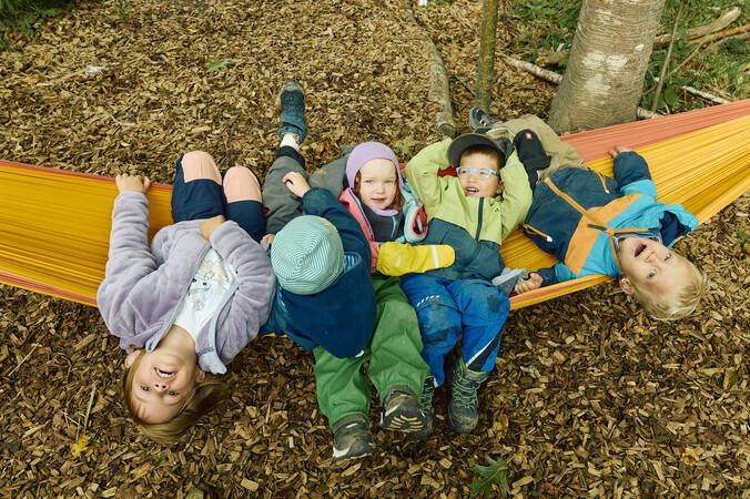 Fünf fröhliche Kinder in bunter Kleidung liegen gemeinsam auf einer bunten Hängematte im Wald neben dem Waldkindergarten.