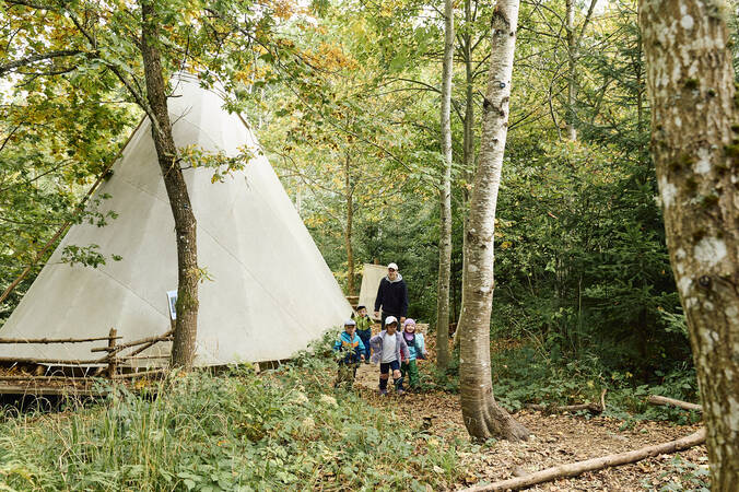 Kinder des Waldkindergartens und ein Erzieher, gehen auf einem Waldpfad vor einem Tipi-Zelt spazieren.