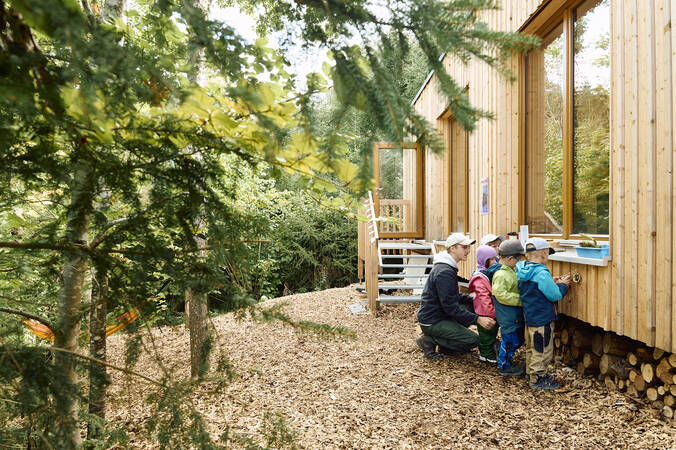 Eine Person und mehrere Kinder erkunden die hölzerne Fassade des Waldkindergartens.