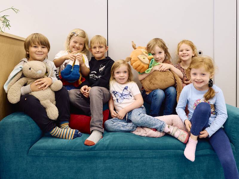 Eine Gruppe von Jungen und Mädchen sitzt lächelnd auf einem blauen Sofa im Kindergarten. Ein paar der Kinder haben ein Kuscheltier in der Hand.