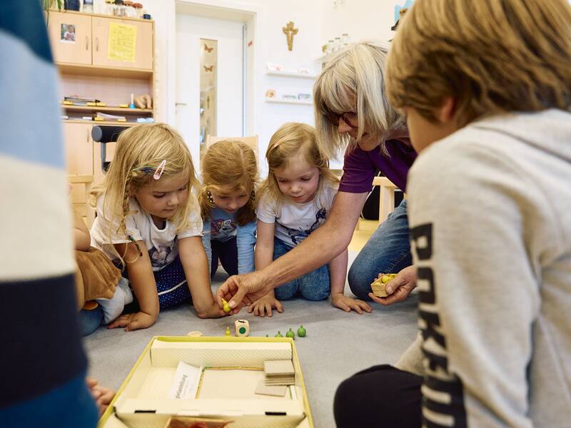 Eine Gruppe von Kindern sitzt auf dem Boden und spielt ein Brettspiel, während eine erwachsene Person ihnen hilft.