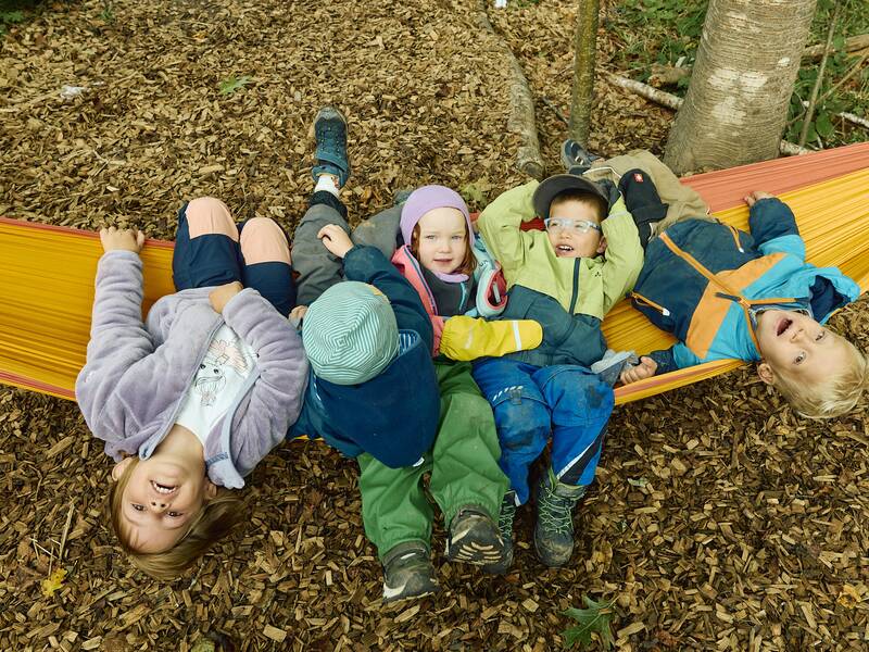 Fünf fröhliche Kinder in bunter Kleidung liegen gemeinsam auf einer bunten Hängematte im Wald neben dem Waldkindergarten.