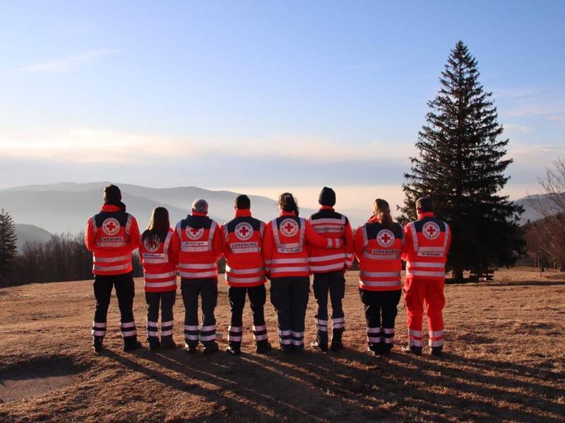 Eine Gruppe von acht Personen in einheitlichen Rotkreuz-Jacken steht mit dem Rücken zur Kamera auf einer Wiese und blickt in eine bergige Landschaft mit Tannenbäumen.