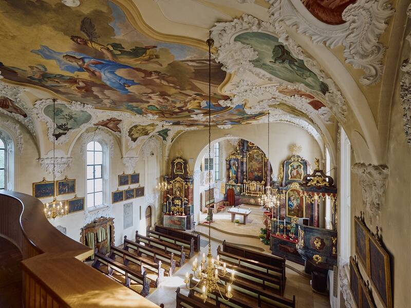 Ein barocker Kirchenraum mit prächtigen Deckenmalereien, vergoldeten Altären und Holzbänken, erhellt von einem Kronleuchter.