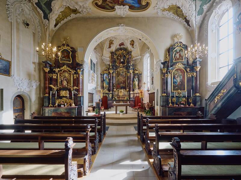 Ein barocker Kirchenraum mit prächtigen Deckenmalereien, vergoldeten Altären und Holzbänken, erhellt von einem Kronleuchter.