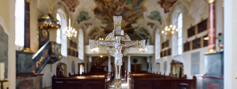 In einer reich verzierten Kirche steht ein kunstvolles Kreuz im Vordergrund auf einem weißen Tuch, während im Hintergrund Bänke, ein Altar und bemalte Decken zu sehen sind.