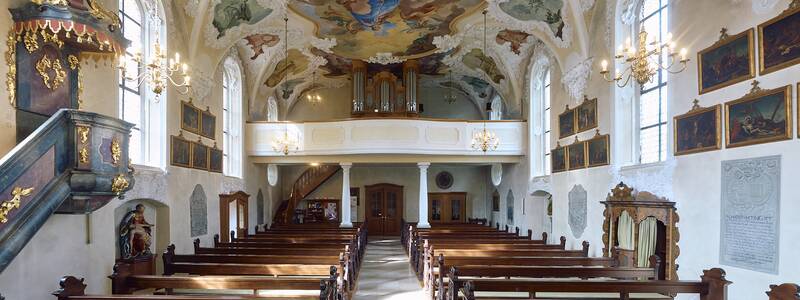 Ein barocker Kirchenraum mit prächtigen Deckenmalereien, vergoldeten Altären und Holzbänken, erhellt von einem Kronleuchter.