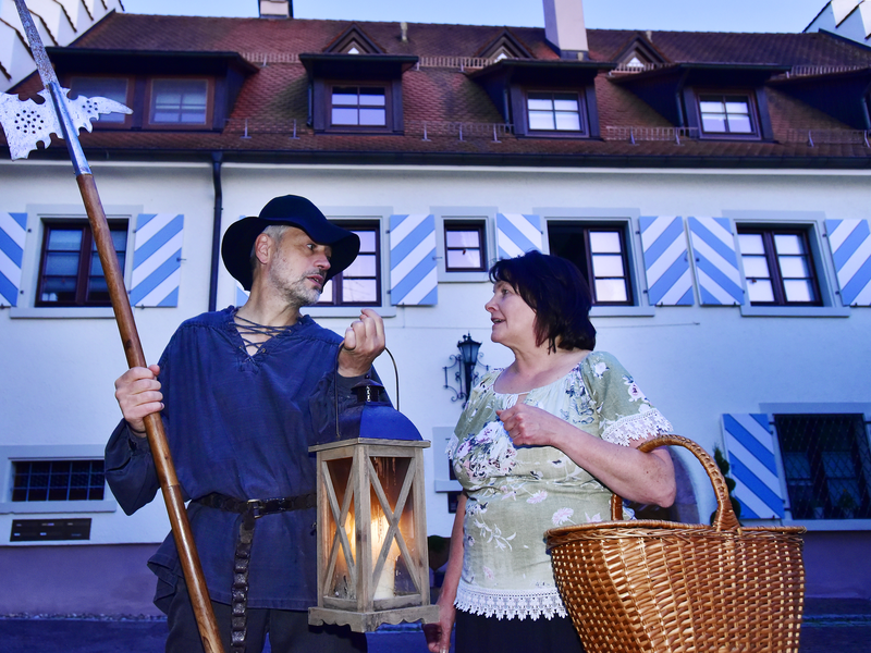 Ein Nachtwächter und eine Hebamme in mittelalterlicher Kleidung und schwarzem Hut hält eine Hellebarde vor einem Fachwerkhaus.