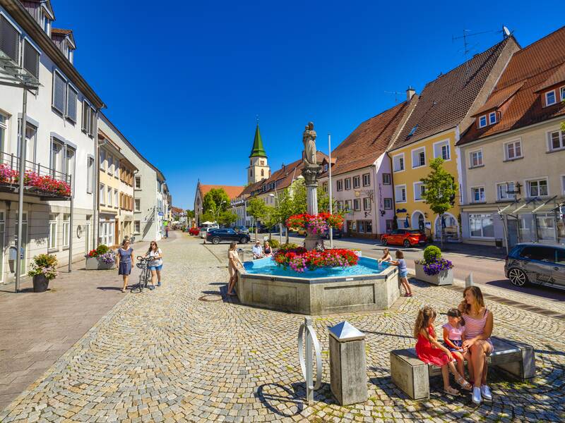 Das Bild zeigt die Innenstadt von Hüfingen, bei dem vorne im Bild eine Frau mit Kind auf einer Bank sitzt. In mitten des Bildes ist ein Brunnen zu sehen und im Hintergrund sind Häuser und eine Kirche zu sehen.
