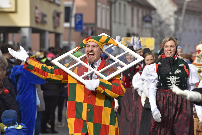 Narrenfigur mit buntem Häs, Fenst trägt er um den Hals 