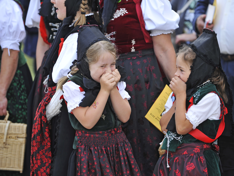 Zwei junge Mädchen in traditionellen Trachtenkleidern stehen lächelnd nebeneinander und halten sich die Hände vor den Mund.