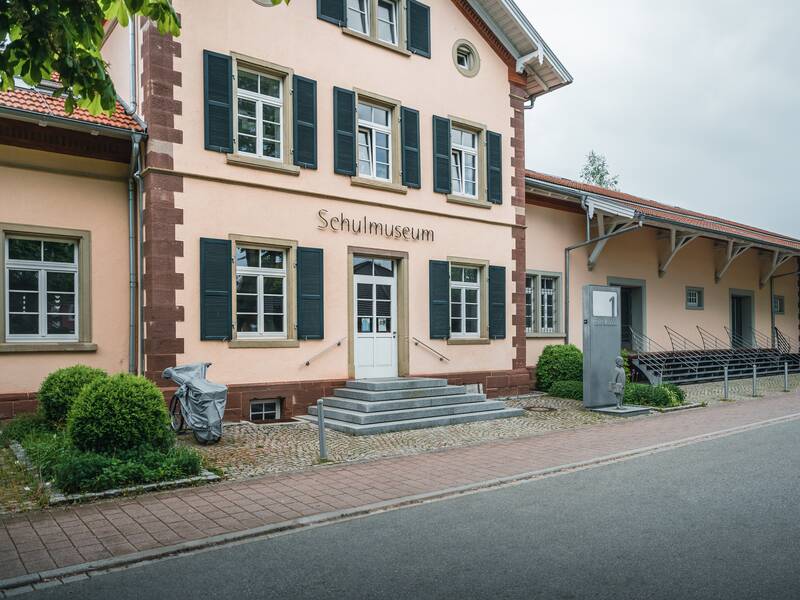 Das Bild zeigt unser Schulmuseum von außen mit cremefarbener Fassade, grünen Fensterläden und der Aufschrift "Schulmuseum".