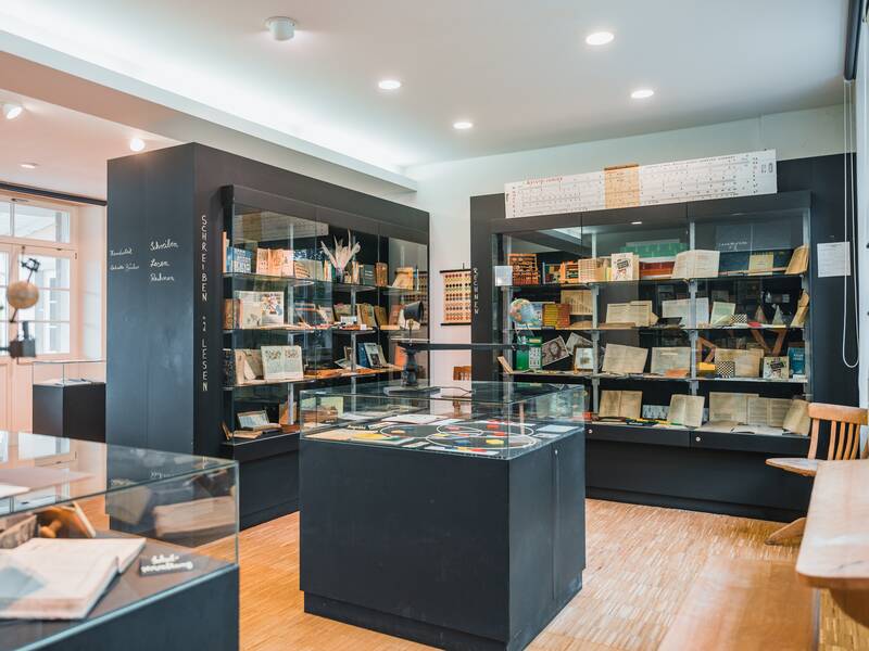 Ein heller Raum mit Glasvitrinen voller historischer Bücher, Dokumente und Lehrmaterialien.