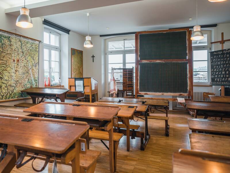 Ein historisches Klassenzimmer mit Holzbänken, einer großen Wandkarte, einer Tafel und einem Abakus.