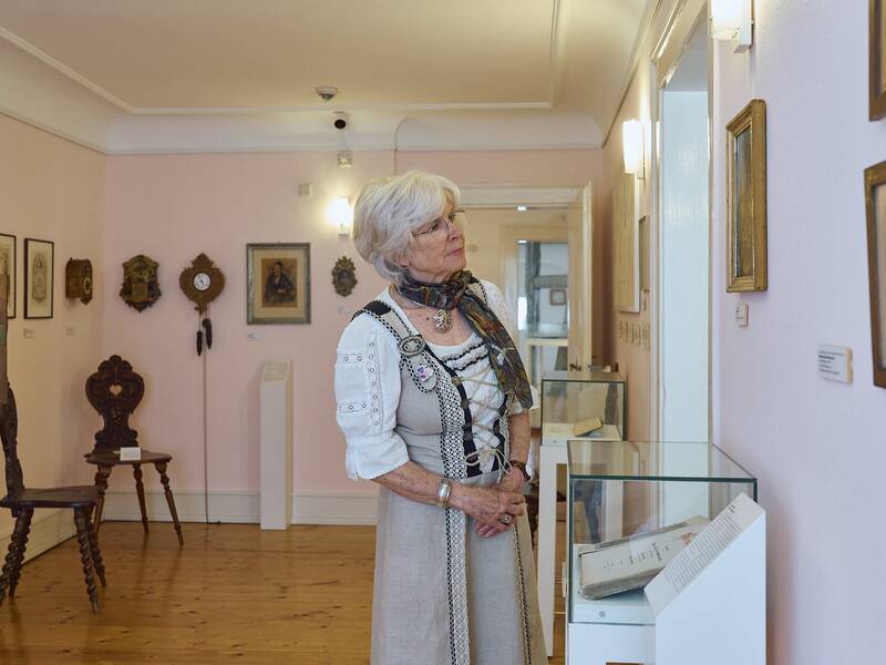 Eine Frau in traditioneller Kleidung betrachtet ausgestellte Kunstwerke im hellen Raum des Stadtmuseums mit Holzboden.