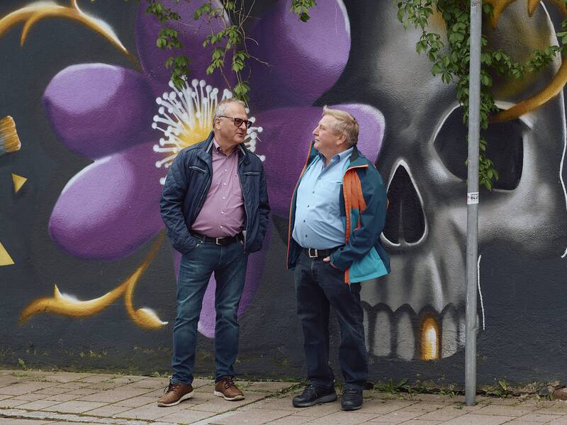 Zwei Männer stehen vor der mit Graffiti bemalten Außenwand des Stadtmuseums, die eine große lila Blume und einen Totenschädel zeigt.