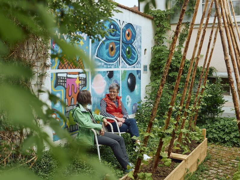 Zwei Personen sitzen im Garten des Stadtmuseums auf Stühlen vor einer bunt bemalten Wand, während Pflanzen an einem Holzgestell hochwachsen.
