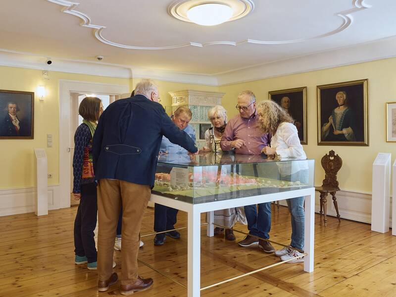 Eine Gruppe von Menschen betrachtet ein Modell der früheren Stadtansicht von Hüfingen in einem Raum im Stadtmuseum mit Holzboden und Porträts an den gelben Wänden.