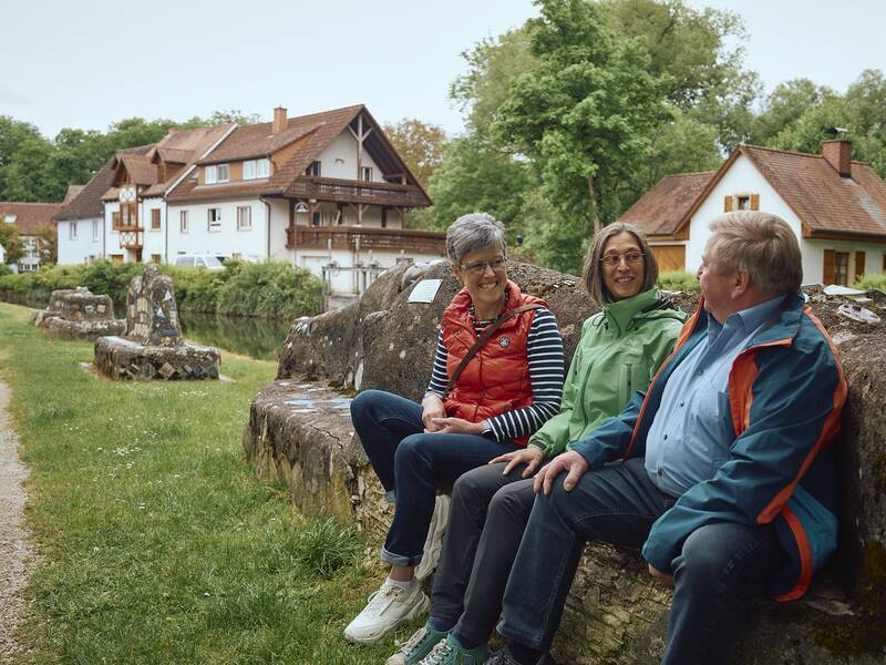 Drei Personen sitzen draußen auf einer Steinmauer und unterhalten sich vor einer ländlichen Kulisse mit Häusern und Bäumen und mit der Breg im Hintergrund.