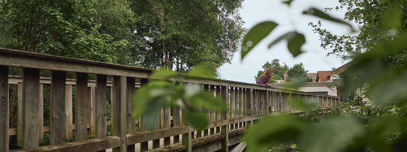 Eine Person beugt sich über eine hölzerne Brücke, die über einen kleinen, von Bäumen umrahmten Fluss führt, und im Vordergrund sind Zweige unscharf zu sehen.