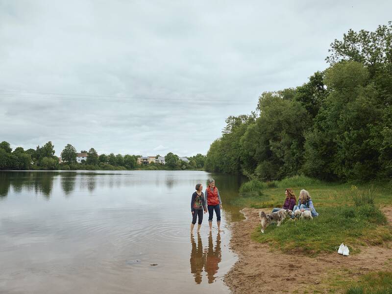 Am Ufer des Kofenweihers stehen zwei Menschen im flachen Wasser, während zwei weitere Personen mit zwei Hunden im Gras sitzen, umgeben von Bäumen.