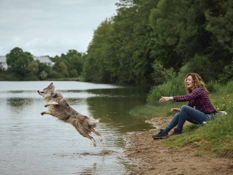 Eine Frau sitzt am Ufer eines Sees und wirft ein Stock, während ein Hund dynamisch ins Wasser springt.
