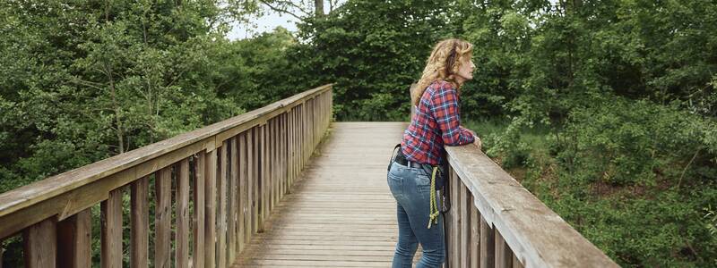 Eine Frau in kariertem Hemd lehnt sich an ein Geländer auf einer Holzbrücke, während ein Hund neben ihr steht und beide auf den dichten grünen Wald blicken.