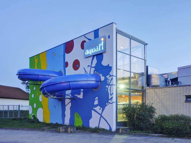 Das Aquari von außen: ein modernes Gebäude mit einem farbenfrohen Wanddesign und zwei blauen Wasserrutschen.