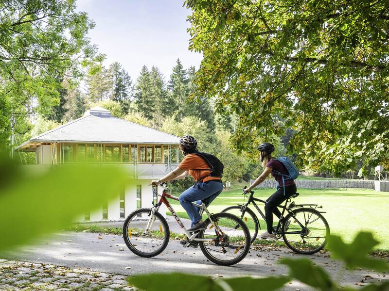 Zwei Menschen fahren mit Fahrrädern auf einem Weg durch eine grüne Parklandschaft. Im Hintergrund ist das Römerbad-Pavillon zu sehen.