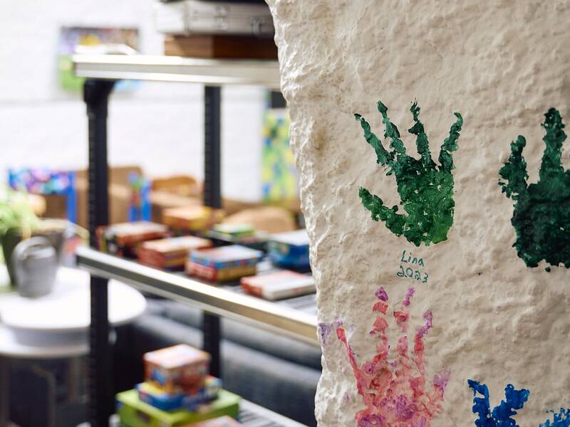 Der rau verputzter Innenwandpfeiler im Jugendhaus ist mit bunten Handabdrücken bemalt, während im Hintergrund mehrere Regale mit Spielen zu sehen sind.
