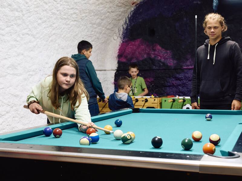 Im Eingangsbereich des Jugendhauses mit bemalter Wand im Hintergrund spielt ein Mädchen Billard, während andere Kinder und ein Jugendlicher im Hintergrund zu sehen sind.