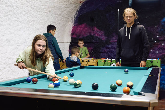 Im Eingangsbereich des Jugendhauses mit bemalter Wand im Hintergrund spielt ein Mädchen Billard, während andere Kinder und ein Jugendlicher im Hintergrund zu sehen sind.