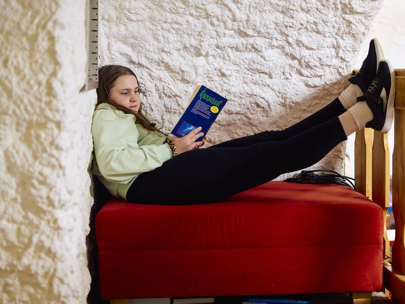 Ein Mädchen liegt entspannt auf einem roten Sofa im Jugendhaus, liest ein Buch und hat ihre Beine auf ein Geländer gelegt.