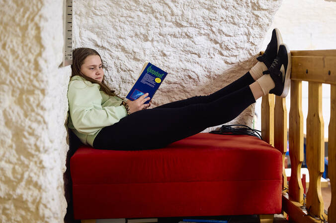 Ein Mädchen liegt entspannt auf einem roten Sofa im Jugendhaus, liest ein Buch und hat ihre Beine auf ein Geländer gelegt.