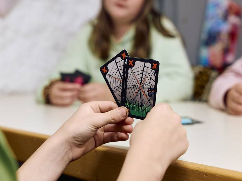 Eine Person hält zwei Spielkarten mit Spinnennetz-Motiven, während andere Personen im Hintergrund ebenfalls Karten in den Händen halten.