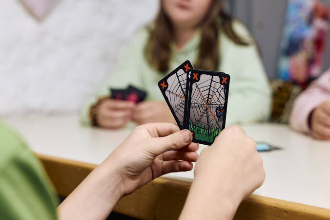Eine Person hält zwei Spielkarten mit Spinnennetz-Motiven, während andere Personen im Hintergrund ebenfalls Karten in den Händen halten.