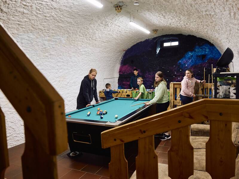 Im Jugendhaus, einem unterirdischen Raum, dessen Wände wie eine Höhle geformt sind, spielen mehrere Personen gleichzeitig an einem Billardtisch und einem Tischkicker.