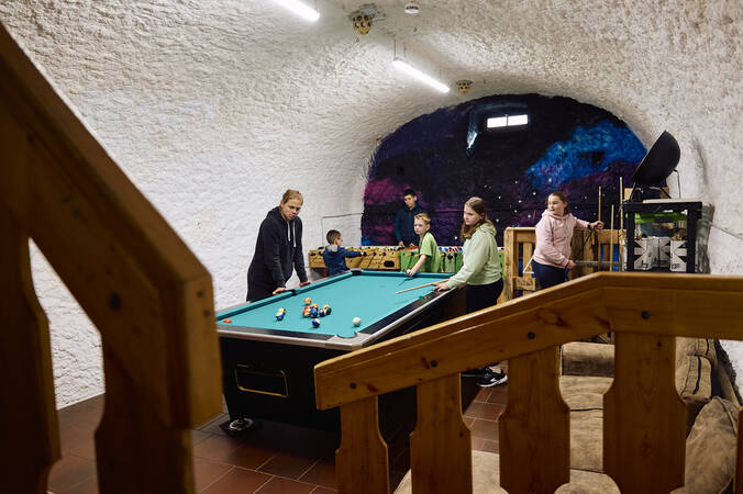 Im Jugendhaus, einem unterirdischen Raum, dessen Wände wie eine Höhle geformt sind, spielen mehrere Personen gleichzeitig an einem Billardtisch und einem Tischkicker.
