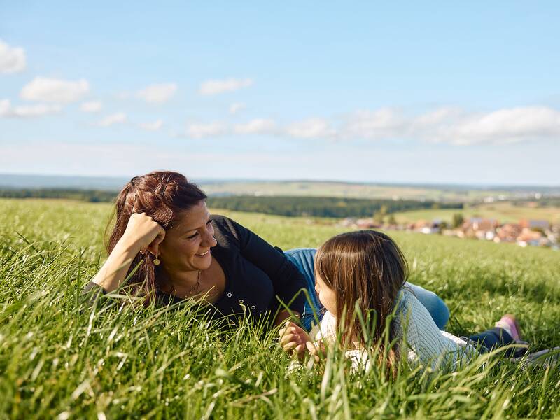 Eine Frau und ein Kind liegen lächelnd im Gras auf einer Wiese mit Blick auf eine weitläufige Landschaft von Behla.