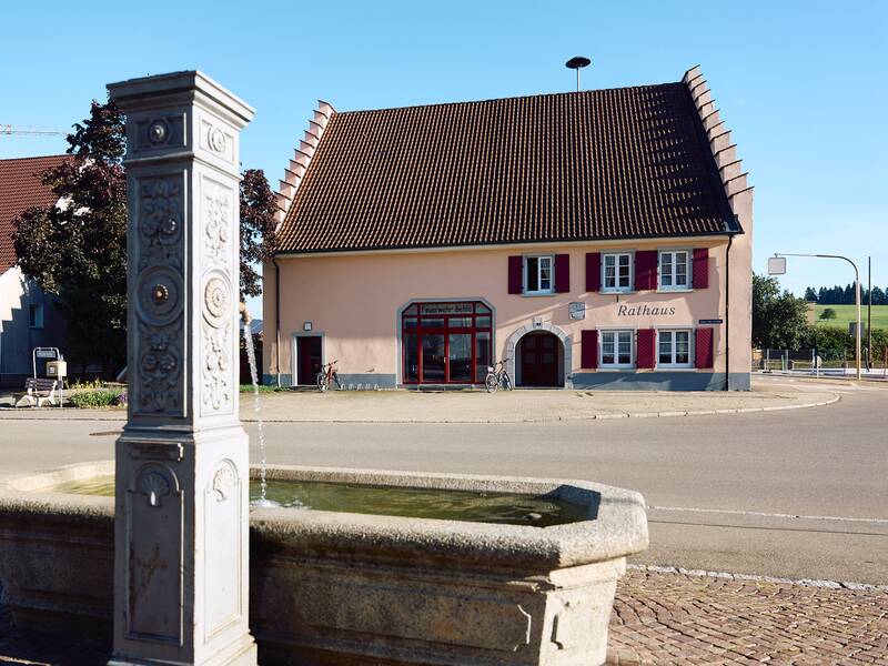 Ein steinerner Brunnen steht im Vordergrund auf einem Platz vor einem rosa Gebäude mit der Aufschrift "Rathaus" und roten Fensterläden.
