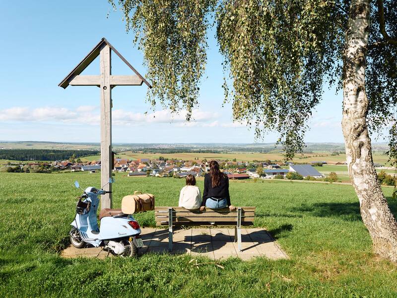 Zwei Personen sitzen auf einer Bank unter einem Baum und schauen auf eine weite Landschaft und Häuser des Ortsteiles Behla, während daneben ein Motorroller und ein großes Holzkreuz stehen.