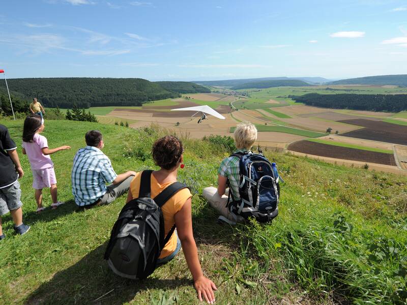 Eine Gruppe von Wanderer beobachtet auf dem Fürstenberg einen Drachenflieger, der sich über einer weiten, grünen Landschaft in die Lüfte erhebt.