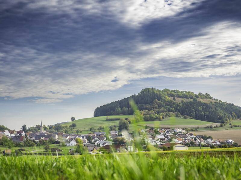 Eine malerische Landschaft des Ortsteils Fürstenberg vor einem bewaldeten Hügel, einem Windrad und einem Sendeturm unter einem dramatisch bewölkten Himmel.