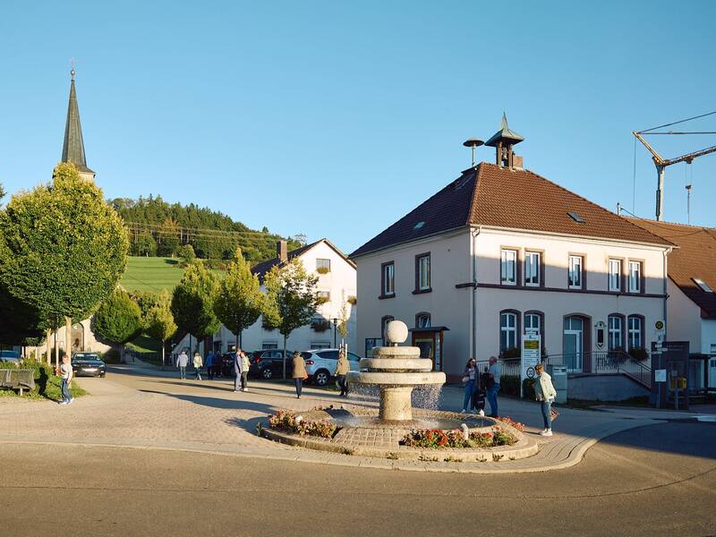 Die Dorfmitte von Fürstenberg mit einem Brunnen in der Mitte, umgeben von Bäumen, einer Kirche im Hintergrund und mehreren Menschen, die spazieren gehen.