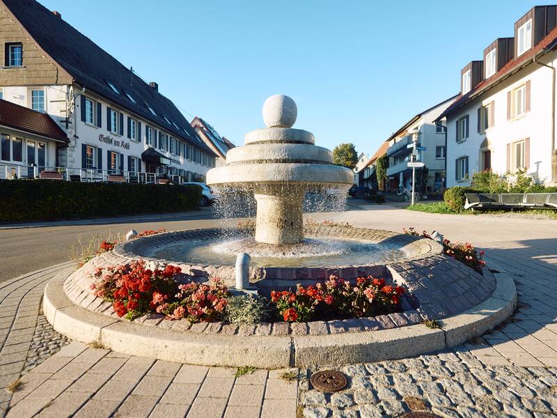 Ein runder Steinbrunnen mit fließendem Wasser steht in einer gepflasterten Straße, umgeben von Blumenbeeten, vor traditionellen Häusern.