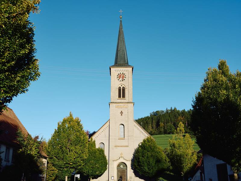 Ein steinerner Kirchturm mit einer Turmuhr und spitzem Dach ragt zwischen grünen Bäumen unter einem klaren blauen Himmel auf.