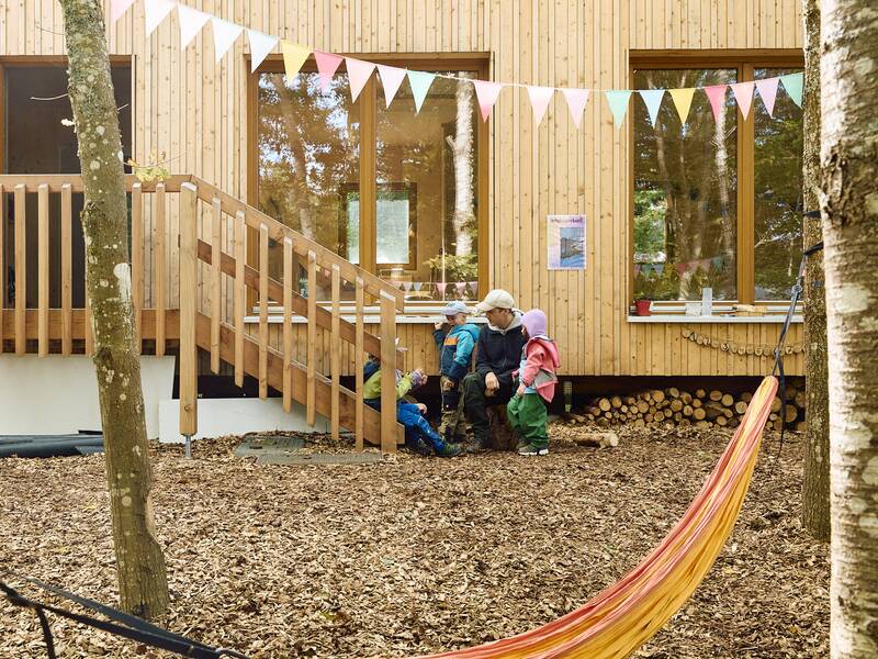 Eine Person und mehrere Kinder erkunden die hölzerne Fassade des Waldkindergartens.