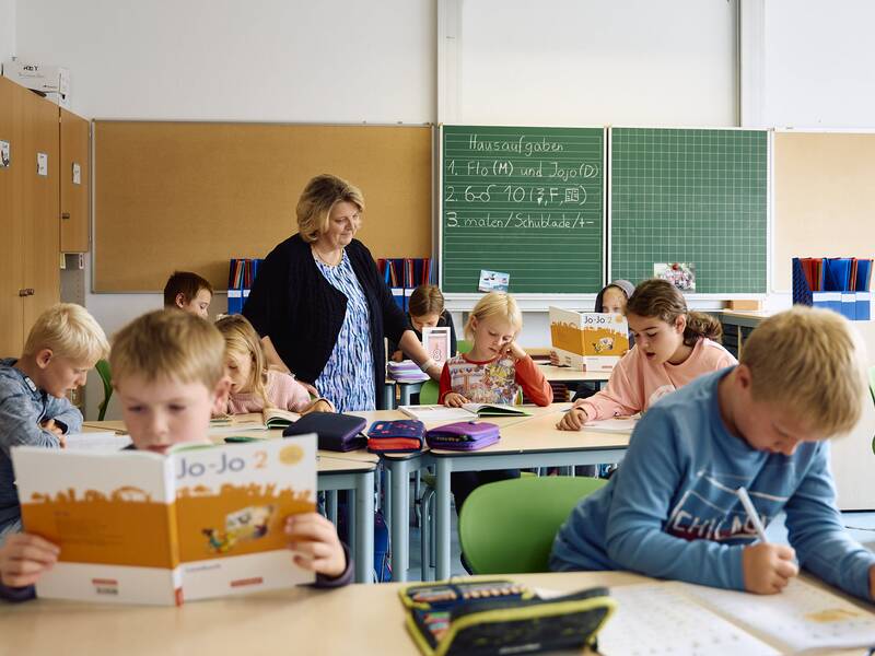 Eine Gruppe von Kindern sitzt an Schultischen im Klassenzimmer der Schellenberger Schule und arbeitet, während eine Lehrerin einem Schüler hilft.